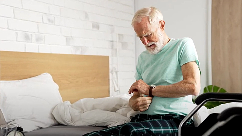 An older man is sitting on the side of a bed putting on his watch.