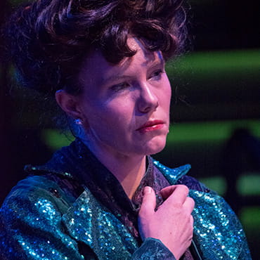 Maggie Whittum, con saco de lentejuelas verde azulado y con expresión de tristeza en el rostro al interpretar una escena de una obra de teatro.