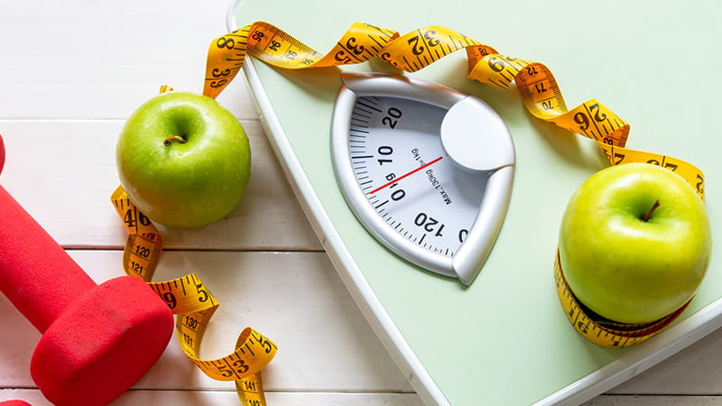 Green apples, a red dumbbell and measuring tape are arranged around a body scale. Green apples, a red dumbbell and measuring tape are arranged around a body scale.
