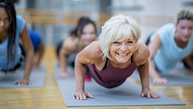 An older White woman is holding a plank pose and smiling while participating in a co-ed, multi-ethnic, fitness class. An older White woman is holding a plank pose and smiling while participating in a co-ed, multi-ethnic, fitness class.