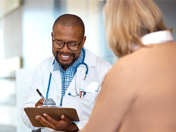 A smiling Black doctor is taking notes while talking with his senior female patient.