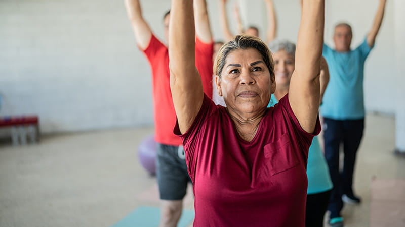 A senior woman is stretching upward with classmates in a yoga studio. A senior woman is stretching upward with classmates in a yoga studio.