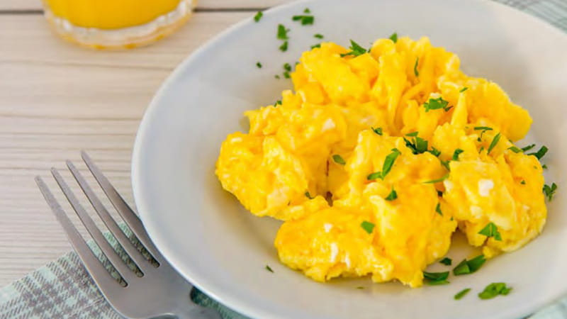 A bowl of scrambled eggs sprinkled with parsley and a fork are on a table. A bowl of scrambled eggs sprinkled with parsley and a fork are on a table.
