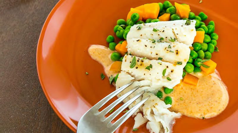 an orange plate of steamed catfish on a bed of peas, carrots and sauce with a fork digging in an orange plate of steamed catfish on a bed of peas, carrots and sauce with a fork digging in