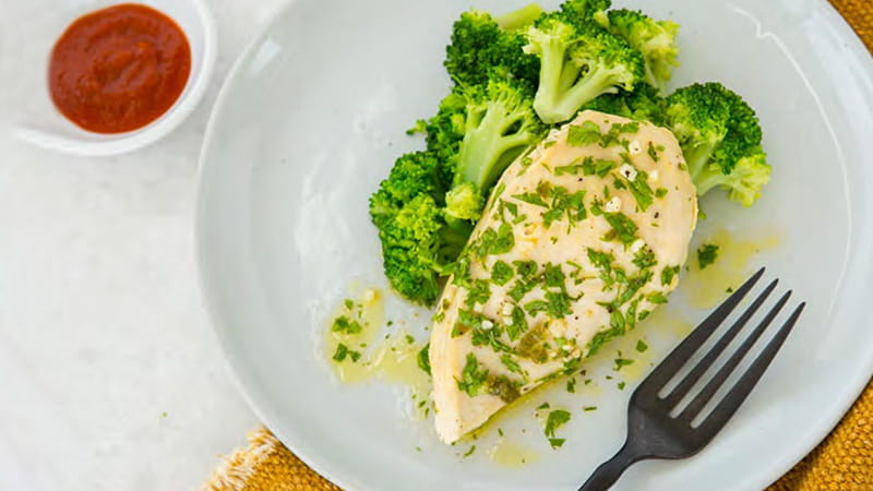 an aerial view of a plate of a steamed chicken breast sprinkled with parsley flakes, broccoli, and a side of sauce an aerial view of a plate of a steamed chicken breast sprinkled with parsley flakes, broccoli, and a side of sauce