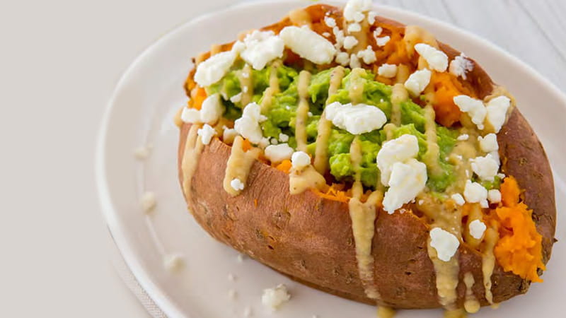 A whole sweet potato stuffed with avocado, feta cheese and drizzled with hummus dressing is on a plate. A whole sweet potato stuffed with avocado, feta cheese and drizzled with hummus dressing is on a plate.