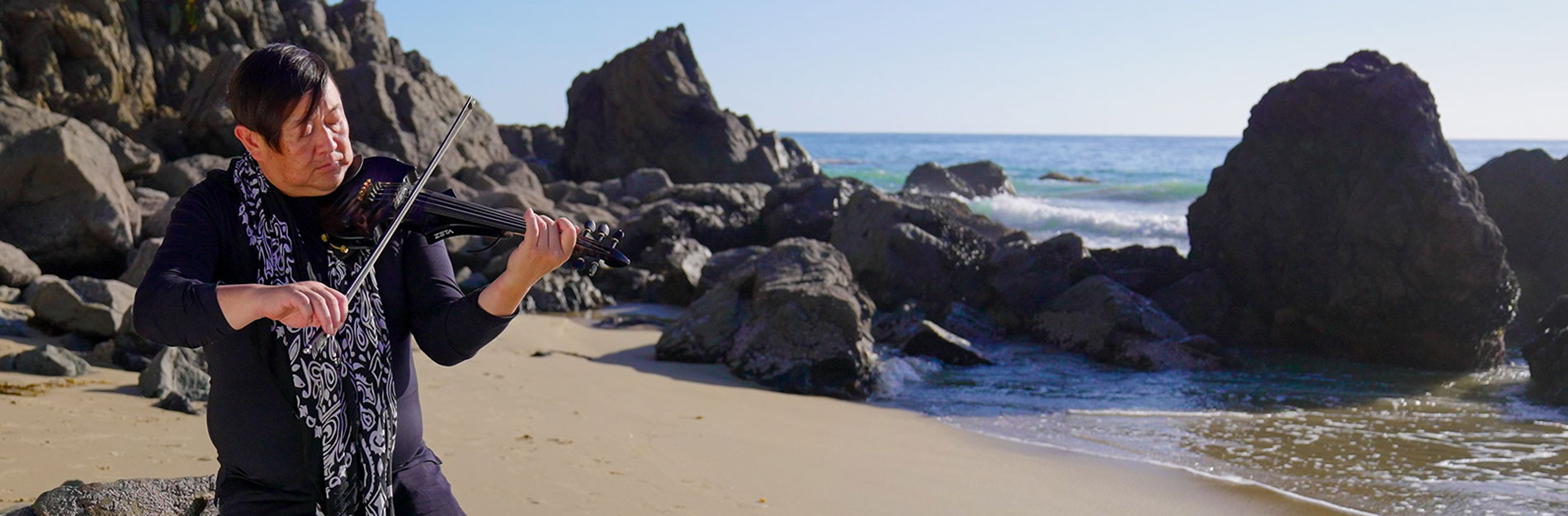 Stroke survivor, Dave, is playing a violin on the beach with the ocean and rocks in the background. Stroke survivor, Dave, is playing a violin on the beach with the ocean and rocks in the background.
