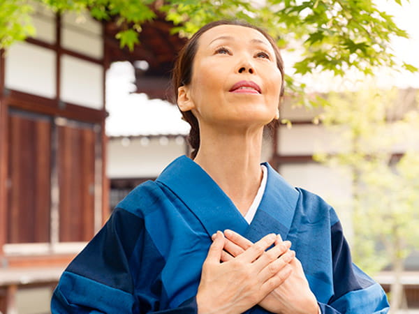 Una mujer japonesa de edad avanzada, vestida con un kimono azul, tiene las manos apoyadas sobre el pecho con un gesto de sanación mientras mira hacia el cielo. Una mujer japonesa de edad avanzada, vestida con un kimono azul, tiene las manos apoyadas sobre el pecho con un gesto de sanación mientras mira hacia el cielo.