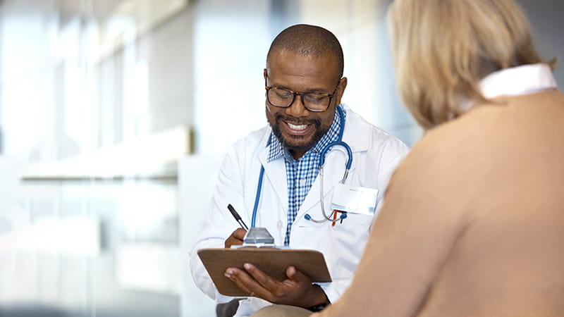 Un médico de raza negra que sonríe toma notas mientras habla con su paciente femenina de edad avanzada. Un médico de raza negra que sonríe toma notas mientras habla con su paciente femenina de edad avanzada.