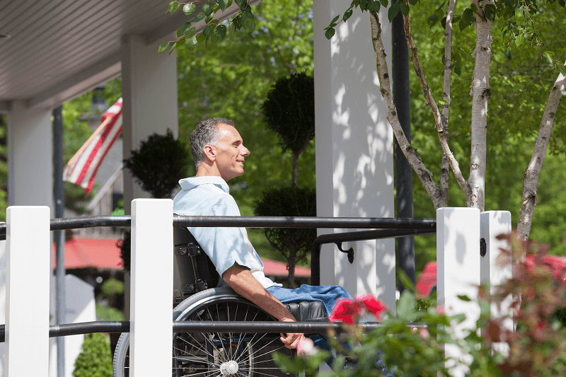 A mature man is sitting in a wheelchair outside on a home porch surrounded by trees. A mature man is sitting in a wheelchair outside on a home porch surrounded by trees.