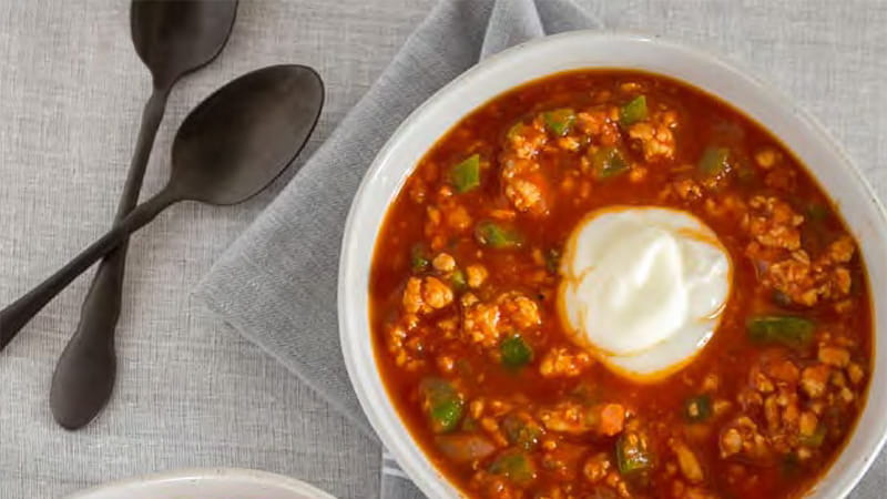 an aerial view of a bowl of chili with a dollop of yogurt and spoons arranged on grey table linens an aerial view of a bowl of chili with a dollop of yogurt and spoons arranged on grey table linens